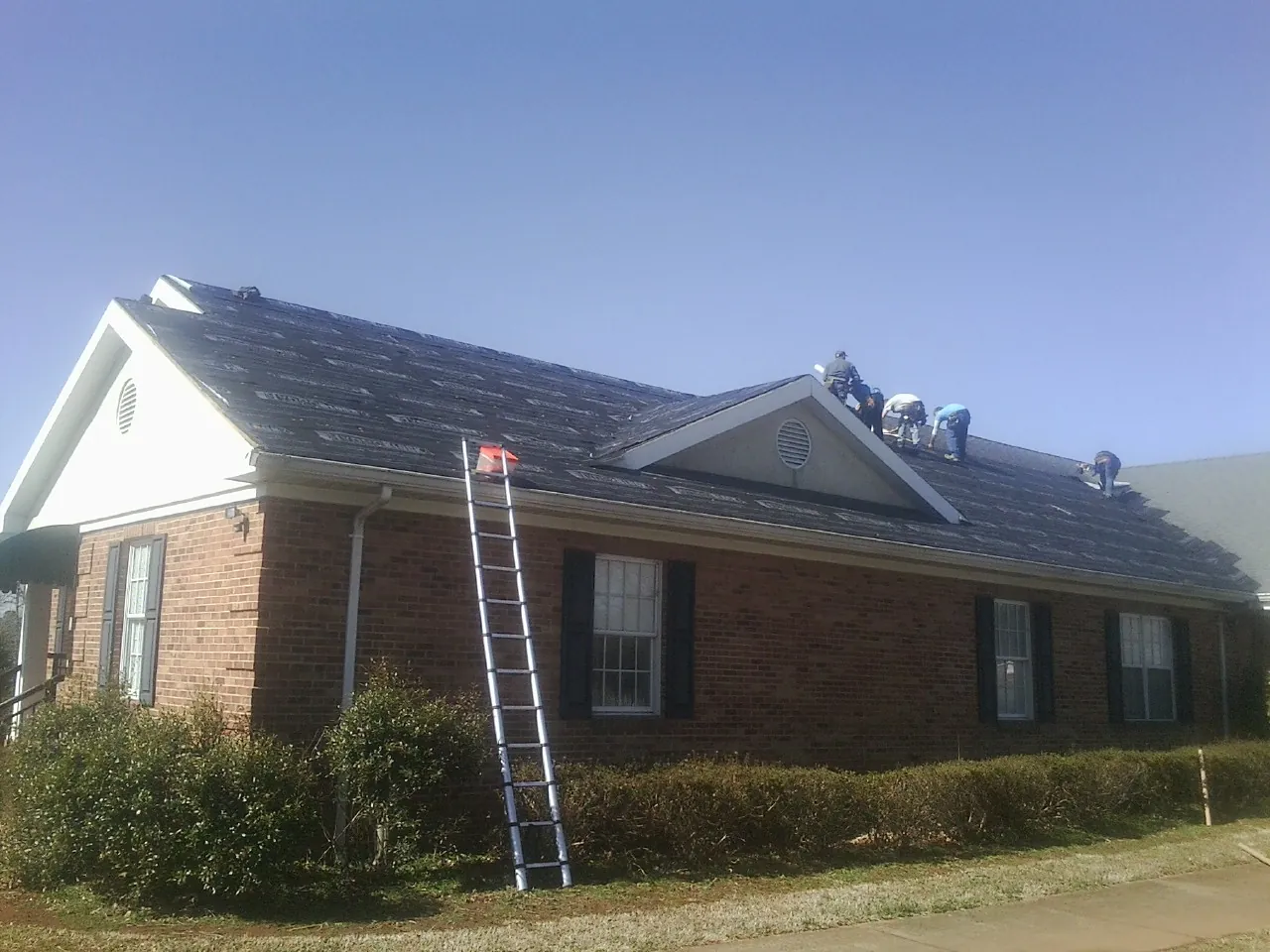 Crew tearing shingles off a brick ranch roof with a ladder set — Metal Roof Repair work by our crew in Cayuga