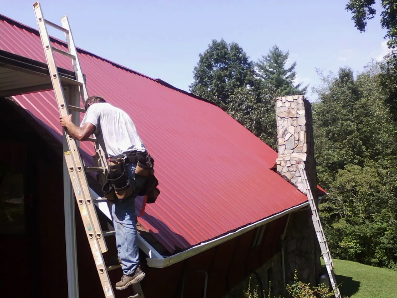 Roofer on a ladder beside a red metal roof and stone chimney — Soffit & Fascia Repair by Roofing Brothers in Cayuga